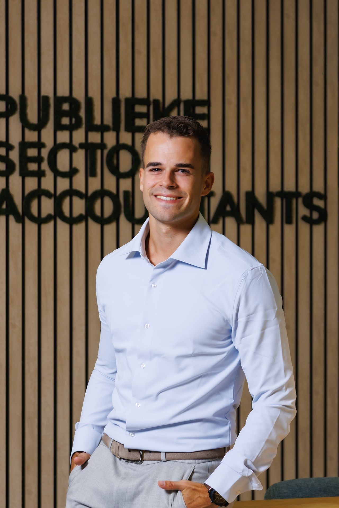 Medewerker van Publieke Sector Accountants poserend voor een houten wand met logo, in een professionele en gastvrije houding.